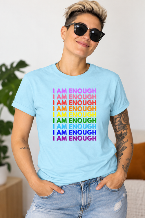A woman wearing a light blue graphic t-shirt with the phrase 'I AM ENOUGH' repeated in stacked rows, each line in a different rainbow color, paired with light-wash distressed jeans and black sunglasses.